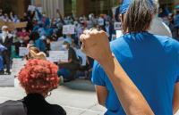 Healthcare workers in masks and scrubs protest, with a person in the foreground raising a fist.